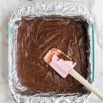 An aluminum foil-lined square pan filled with brownie batter is being spread with a pink spatula on a marble surface.