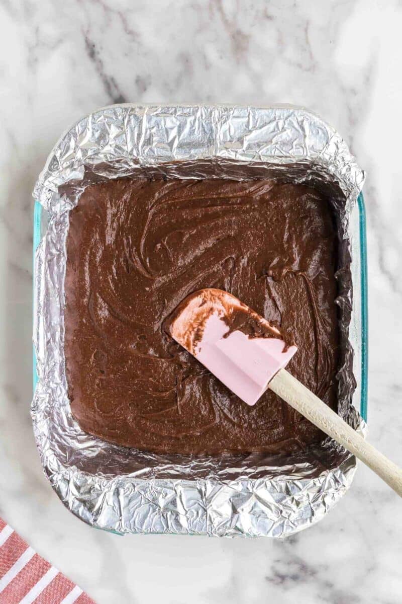 An aluminum foil-lined square pan filled with brownie batter is being spread with a pink spatula on a marble surface.