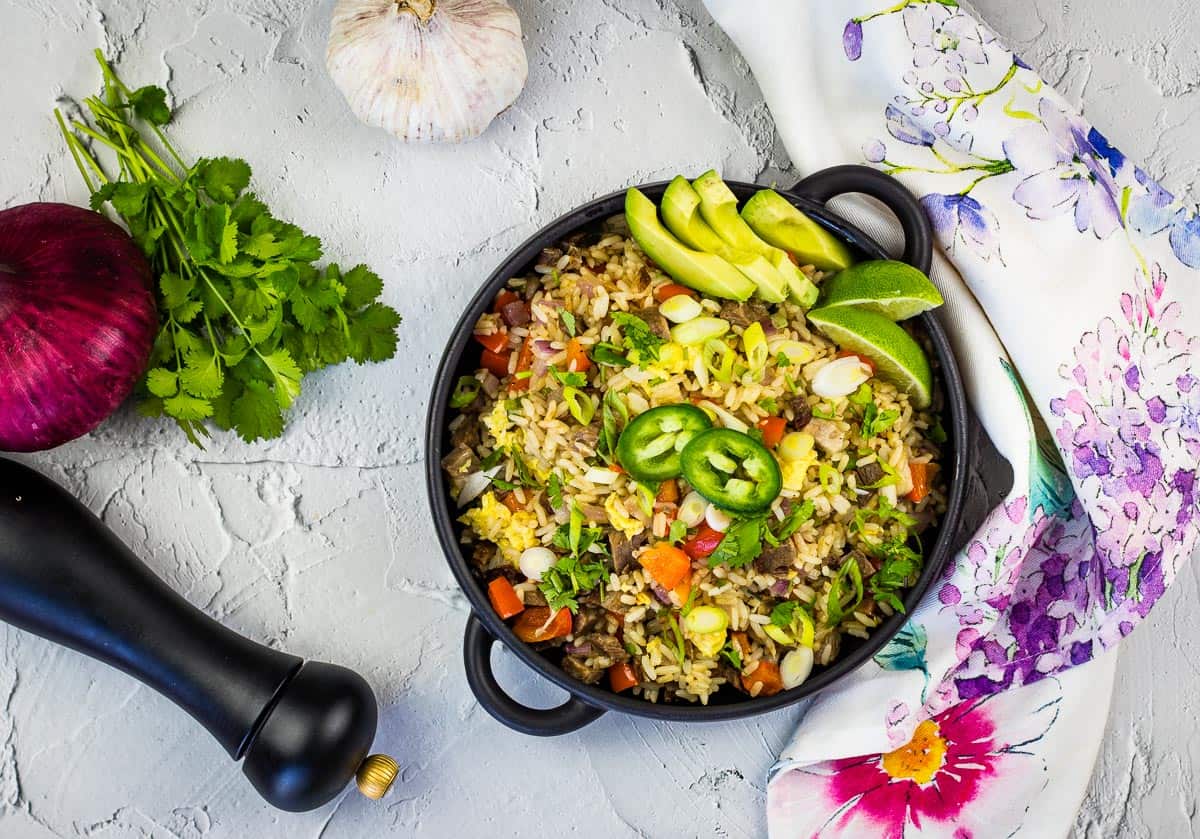 A bowl of easy fried rice garnished with avocado slices, lime wedges, and jalapeño, surrounded by cilantro, red onion, garlic, a pepper grinder, and a floral napkin delivers homemade comfort with takeout flavor.
