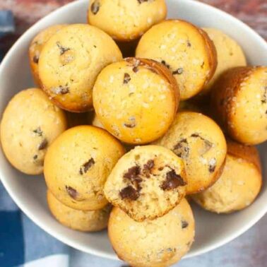 A white bowl filled with chocolate chip muffins, some topped with coarse sugar and boasting tangy crusts and chewy centers, sits beside a blue checked napkin with crumbs visible nearby.