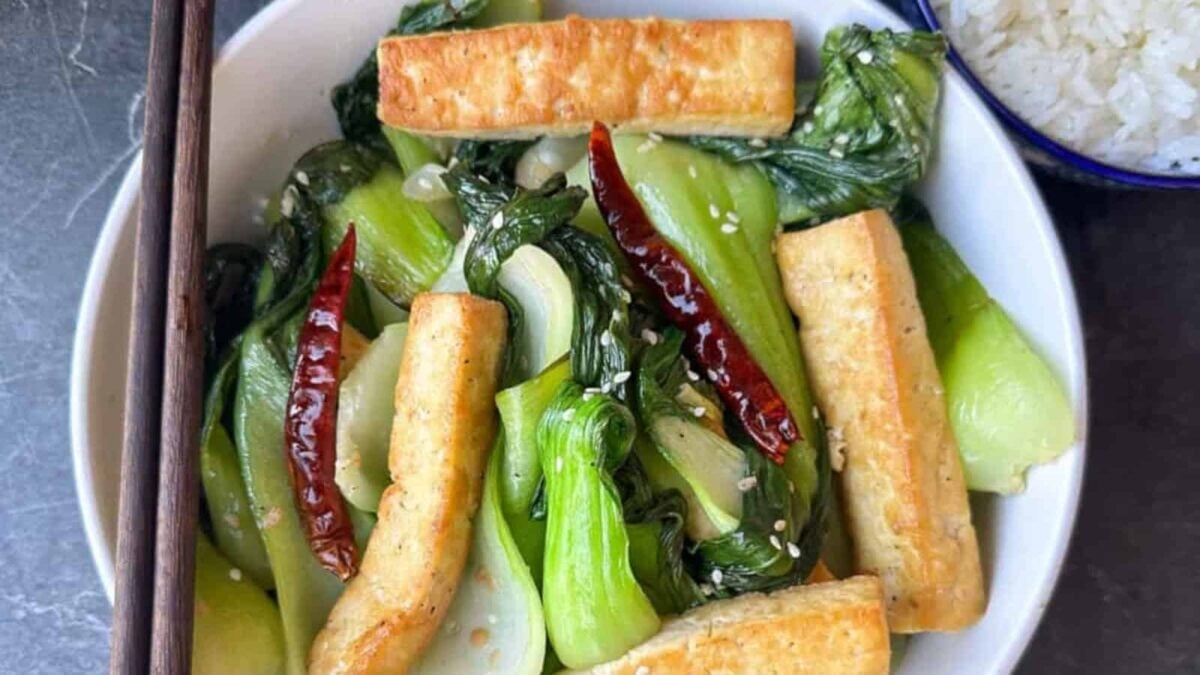 A bowl of stir-fried bok choy with tofu strips boasting crispy edges, dried red chilies, and sesame seeds, served with white rice and chopsticks on the side.