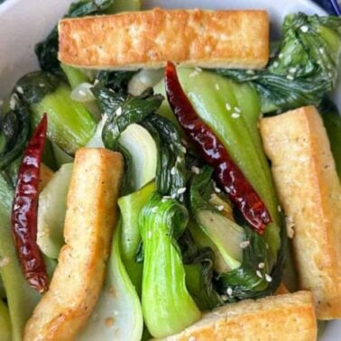 A bowl of stir-fried bok choy with tofu strips boasting crispy edges, dried red chilies, and sesame seeds, served with white rice and chopsticks on the side.