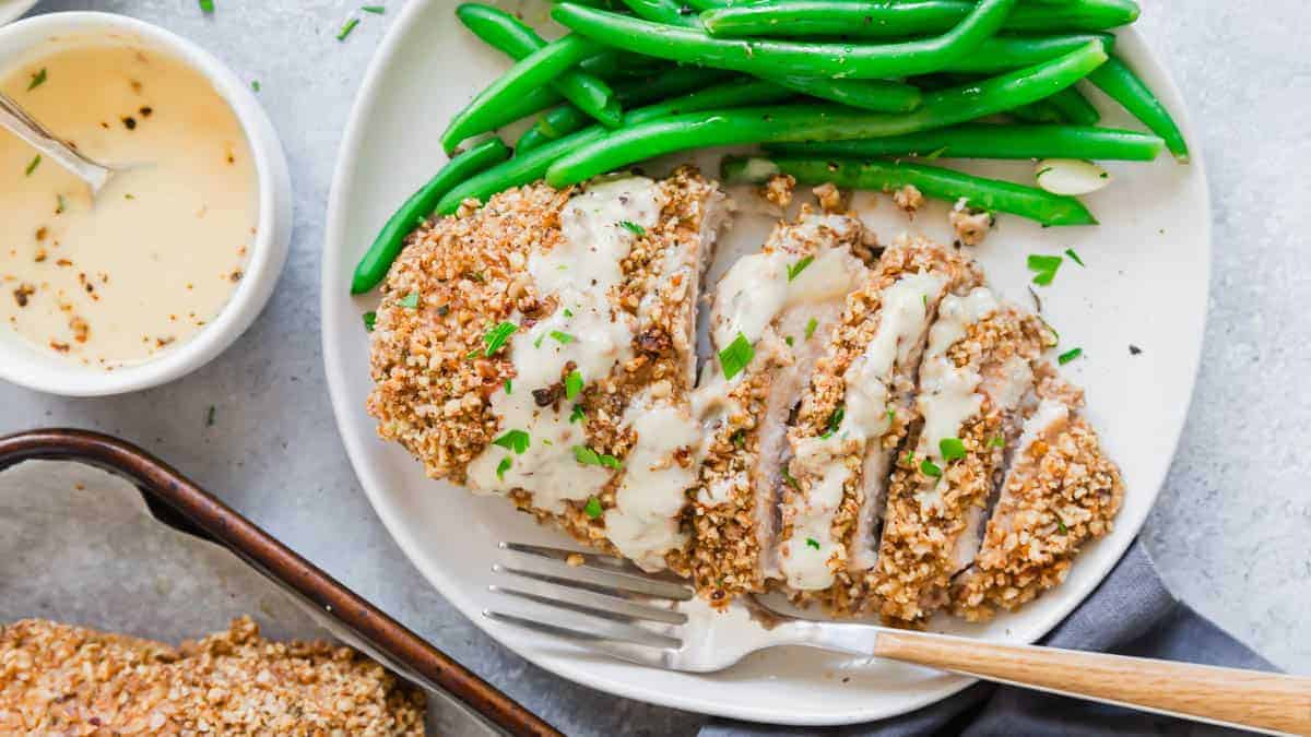 A plate with sliced crunchy chicken topped with creamy sauce, green beans on the side, and a fork; sauce in a small bowl nearby—perfect for fans of crispy chicken recipes.