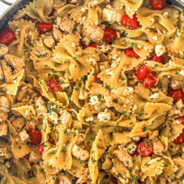 A skillet filled with bowtie pasta, diced chicken for extra protein, cherry tomatoes, crumbled cheese, and herbs.