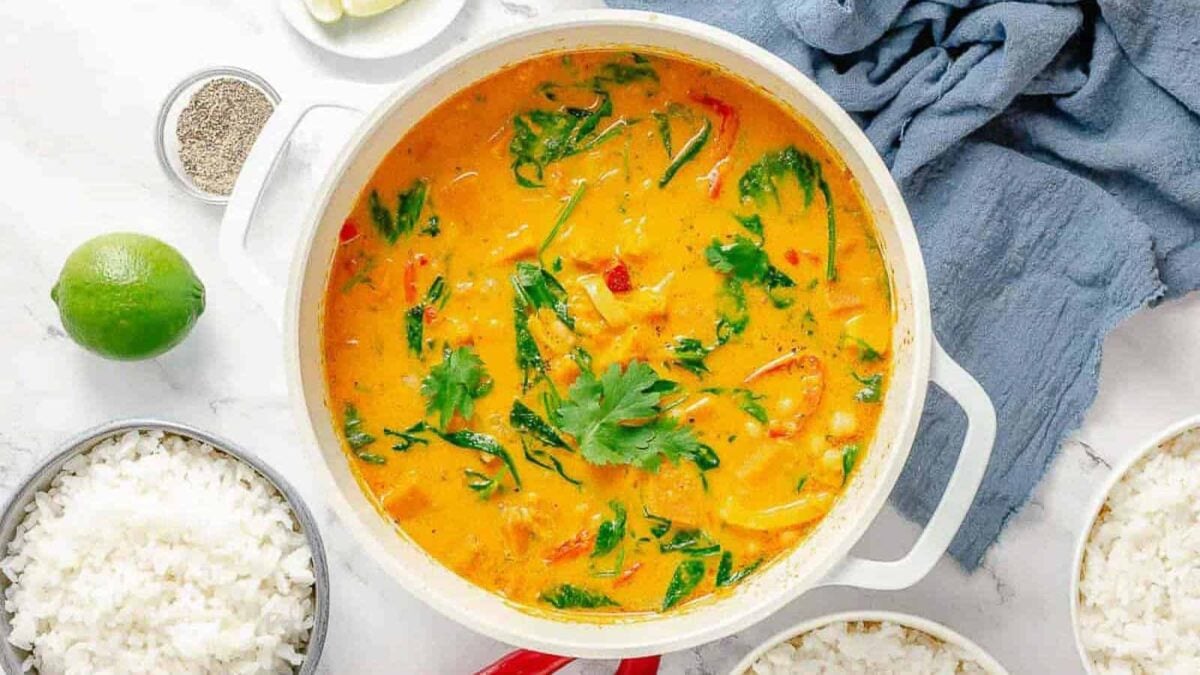 A pot of creamy orange curry with vegetables and greens, rich in warm spices and deep flavor, is garnished with cilantro and sits on a white surface next to bowls of rice, lime, pepper, and a blue cloth.