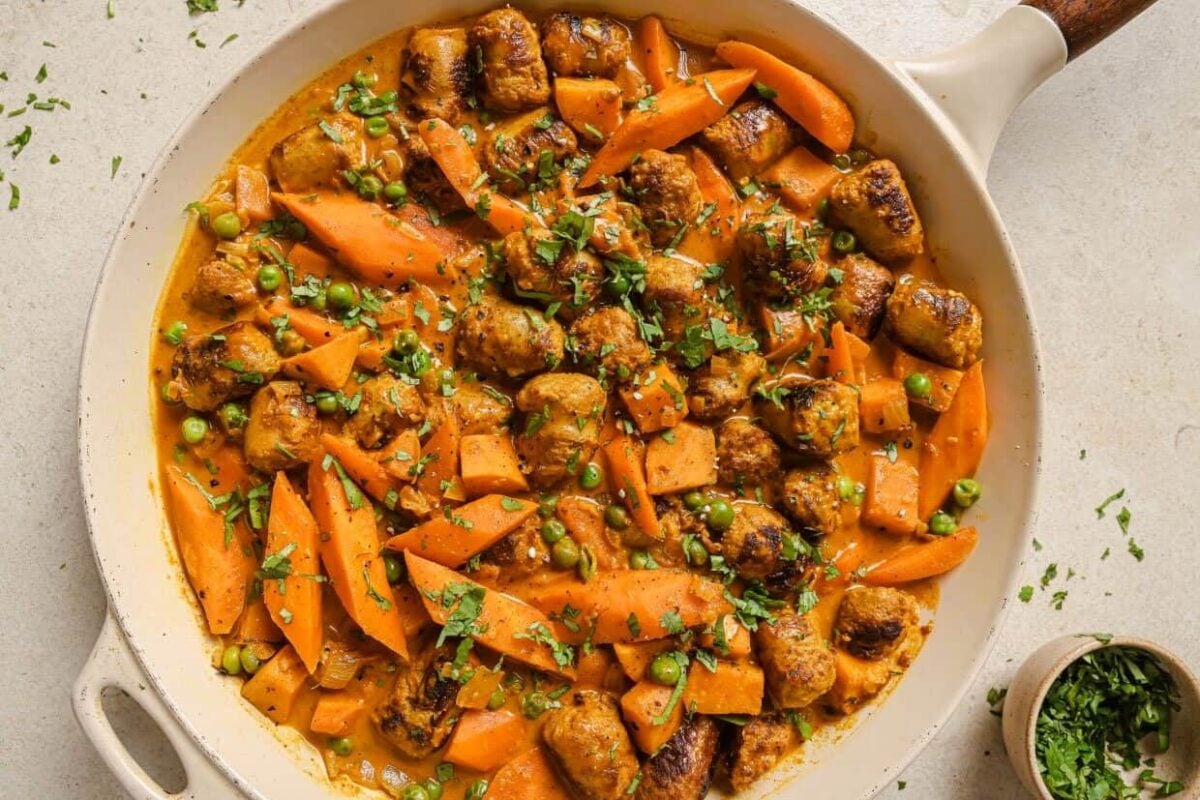 A skillet filled with cooked sausage pieces, sliced carrots, peas, and a creamy orange sauce infused with warm spices, garnished with chopped herbs.