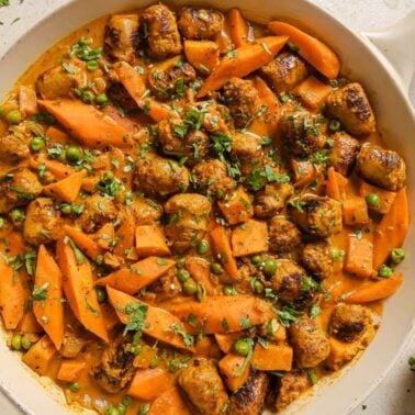 A skillet filled with cooked sausage pieces, sliced carrots, peas, and a creamy orange sauce infused with warm spices, garnished with chopped herbs.