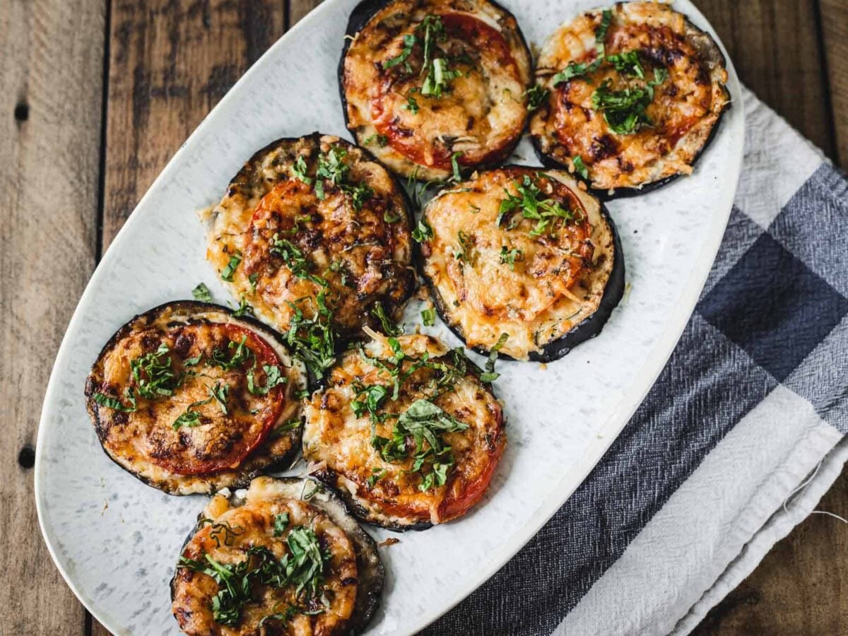 A white oval plate with seven baked eggplant slices topped with melted cheese, tomato, and chopped herbs brings fresh kitchen flavor to your table, perfect for those who love bright and saucy tomato recipes.