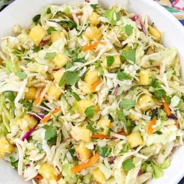 A bowl of coleslaw with shredded cabbage, carrots, pineapple chunks, cilantro, and a light dressing sits on a colorful tableclothโperfect for cozy meals or anyone craving a fresh bite of classic cabbage recipes.