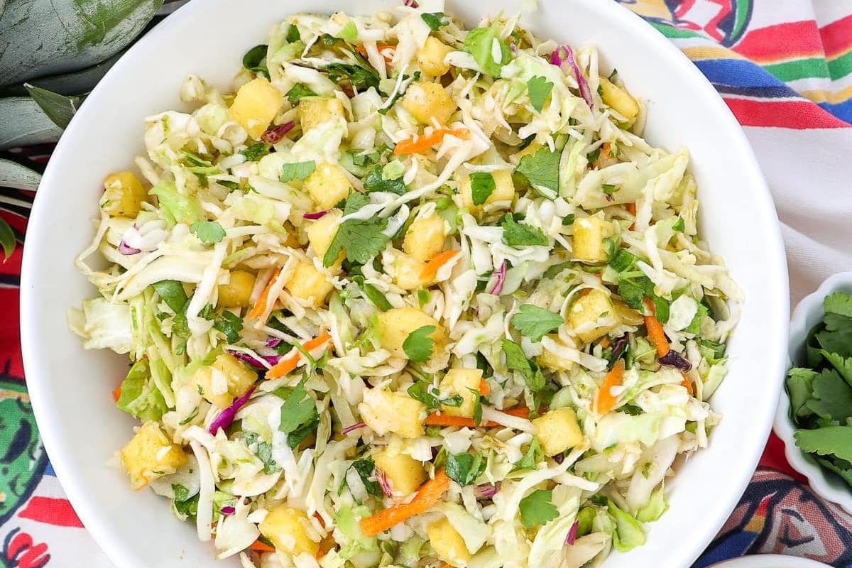 A bowl of coleslaw with shredded cabbage, carrots, pineapple chunks, cilantro, and a light dressing sits on a colorful tablecloth—perfect for cozy meals or anyone craving a fresh bite of classic cabbage recipes.