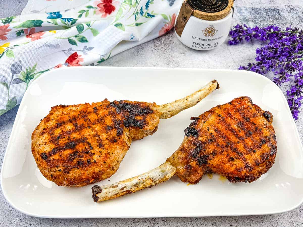 Two grilled pork chops with grill marks on a white rectangular plate, perfect for Low Carb Dinner ideas. Placed on a light surface with a floral napkin, spice jar, and purple flowers nearby.