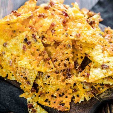 A pile of crunchy, baked cheese chips with browned spots sits on a wooden serving board atop a dark clothโan irresistible, snackable chips delight.