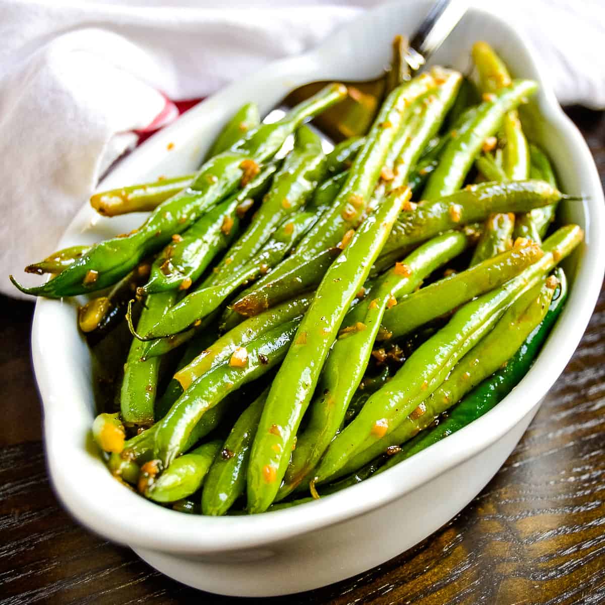 A white oval dish filled with sautéed fresh green beans, garnished with minced garlic and seasonings, rests on a dark wooden surface, showcasing bright vegetables.