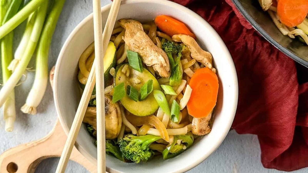 A bowl of stir-fried noodles with chicken, broccoli, zucchini, carrots, and chopped green onions—one of our bold recipes—served with chopsticks resting on top.