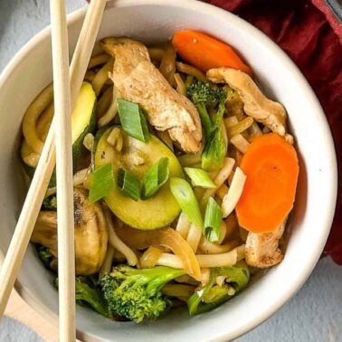 A bowl of stir-fried noodles with chicken, broccoli, zucchini, carrots, and chopped green onionsโone of our bold recipesโserved with chopsticks resting on top.