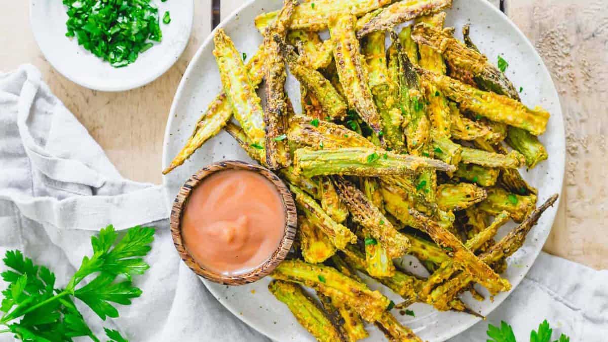 A plate of crispy okra fries, perfect for snack time, is served with a small bowl of dipping sauce and garnished with chopped parsley on a light-colored surface.