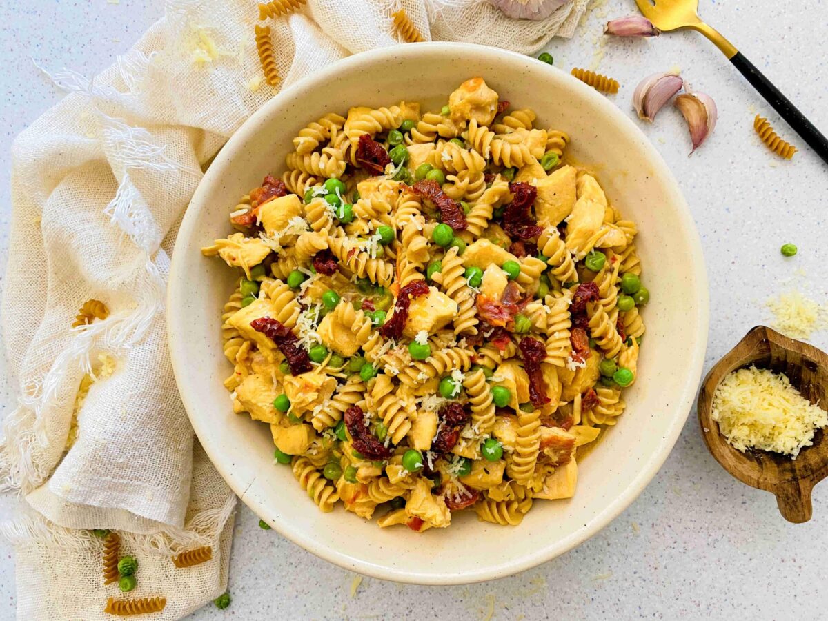 A bowl of rotini pasta with crockpot chicken, peas, sun-dried tomatoes, and grated cheese on a white surface, surrounded by loose pasta and kitchen utensils—perfect inspiration for easy dinner recipes.