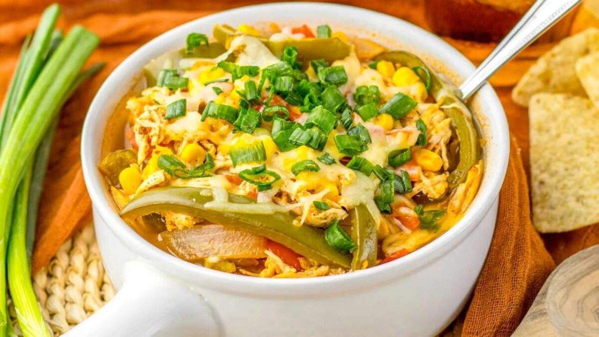 A white bowl filled with a baked chicken casserole—perfect for dinner—topped with melted cheese, sliced green onions, corn, and bell peppers, with a spoon in the bowl and tortilla chips nearby. Great for fans of slow cooker chicken recipes.