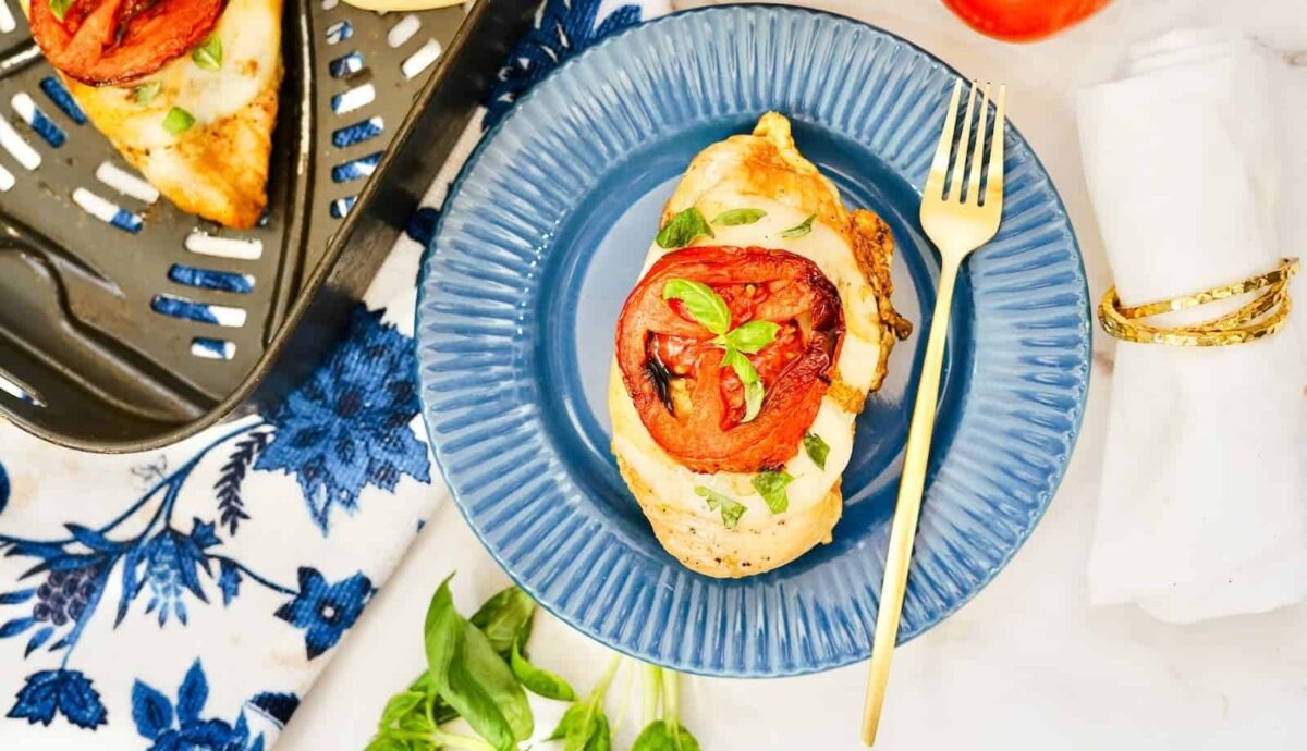 A cooked chicken breast topped with melted cheese, basil, and a roasted tomato slice delivers crispy comfort—served on a blue plate with a gold fork, it's the perfect simple dinner.