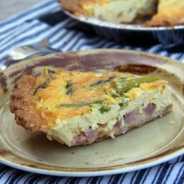 A slice of quiche with ham, cheese, green vegetables, and smoky bacon on a ceramic plate, placed on a striped cloth with a fork beside it.