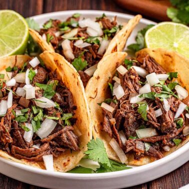 Three tacos filled with slow cooker beef, chopped onions, and cilantro are served on a plate with lime halves, set on a wooden tableโperfect for those craving tender bites.