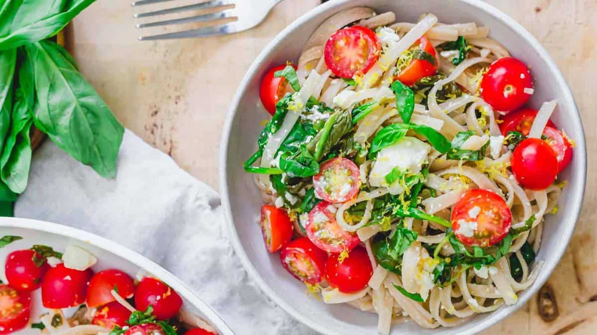 A bowl of pasta topped with halved cherry tomatoes, fresh basil, grated cheese, and a fork placed next to the bowl highlights fresh color and a savory taste.