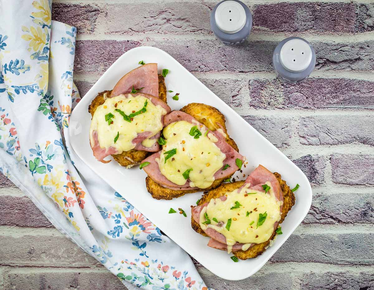 A white platter with four slices of bread topped with ham, melted cheese, and chopped herbs, placed on a patterned cloth next to salt and pepper shakers—perfect for easy weeknight dinners.