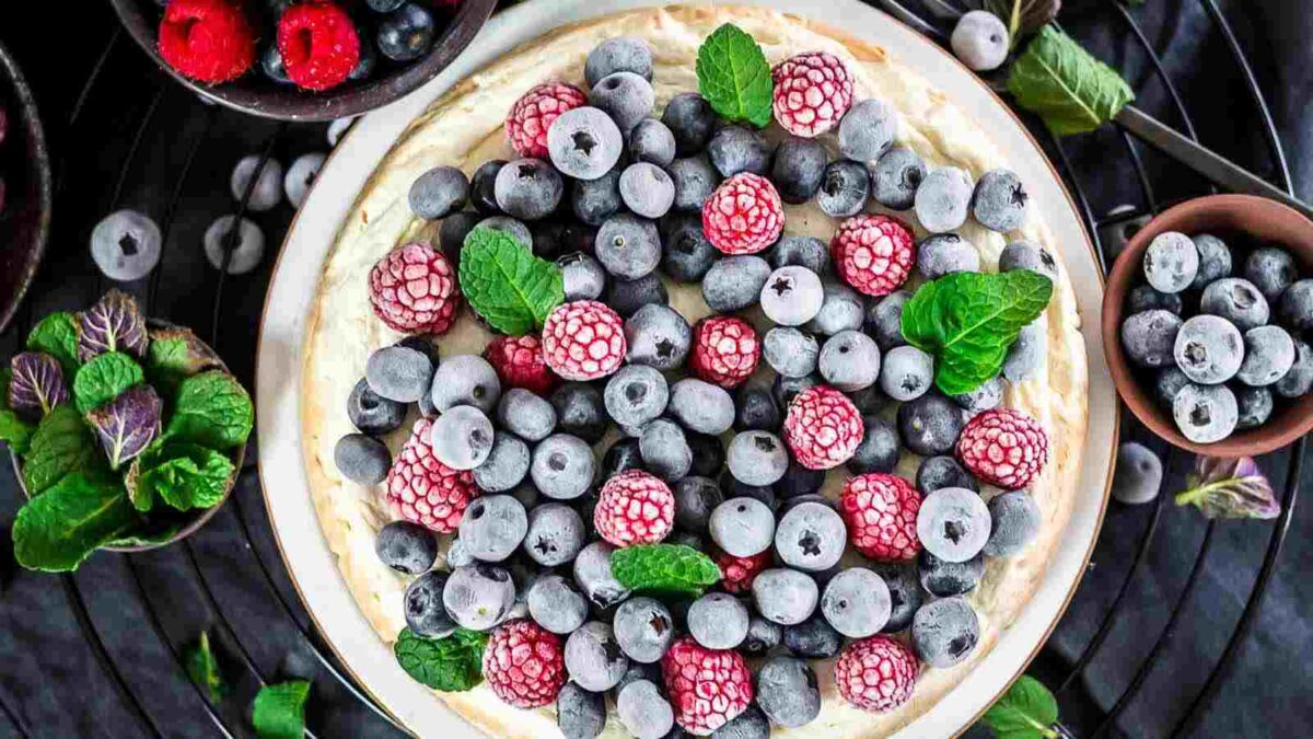 A creamy cheesecake topped with frozen blueberries, raspberries, and fresh mint leaves, surrounded by bowls of berries and mint on a dark surface.