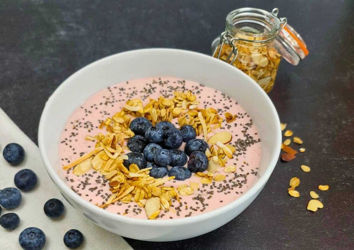 A bowl of creamy pink smoothie topped with blueberries, sliced almonds, and chia seeds sits alongside a jar of sliced almonds and loose blueberries on a dark surface.