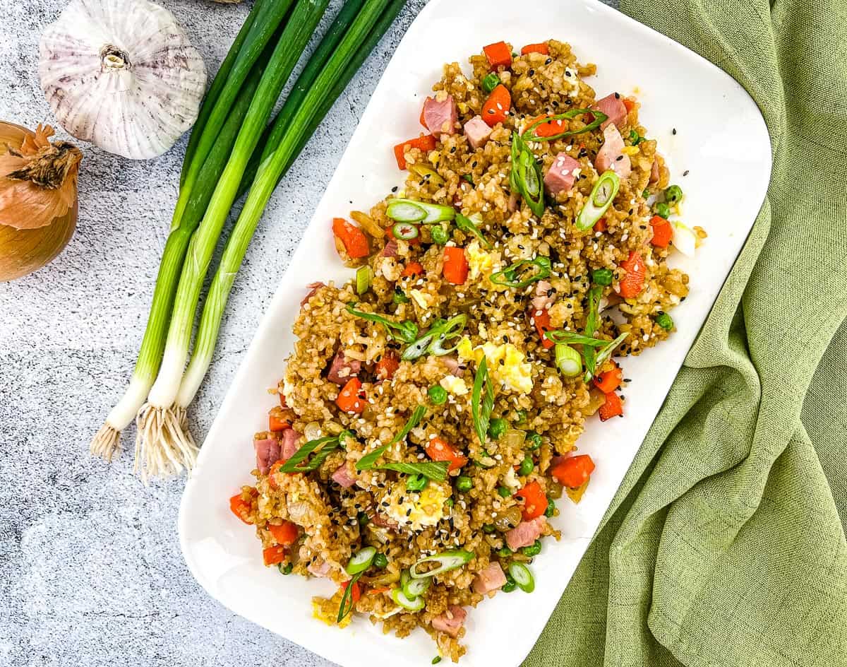 A rectangular white plate with easy fried rice recipes featuring diced ham, carrots, scrambled egg, and green onions, served next to whole green onions, garlic, and an onion on a gray surface for that classic takeout flavor.