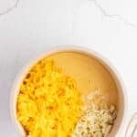 A saucepan containing shredded cheddar and mozzarella cheeses partially melted, sitting on a light-colored surface with a whisk nearby.