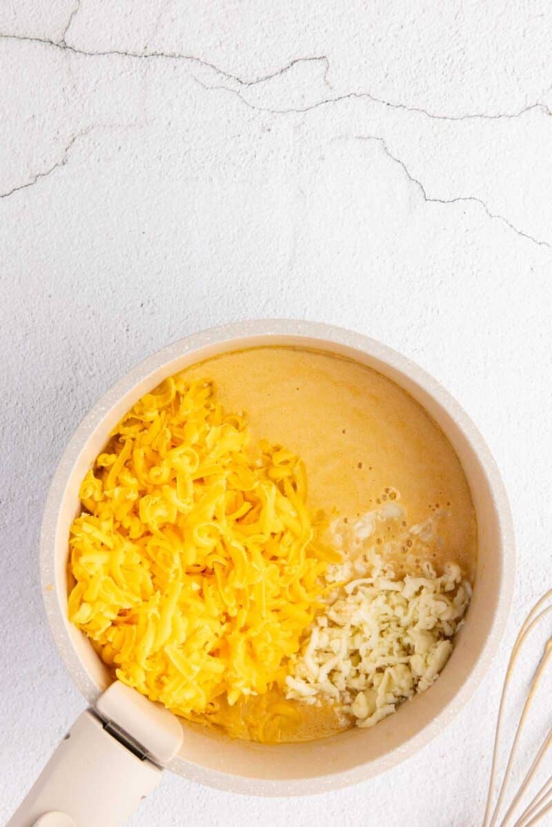 A saucepan containing shredded cheddar and mozzarella cheeses partially melted, sitting on a light-colored surface with a whisk nearby.