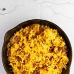 A cast iron skillet filled with macaroni and cheese topped with shredded cheddar cheese and bacon pieces, set on a light-colored surface.
