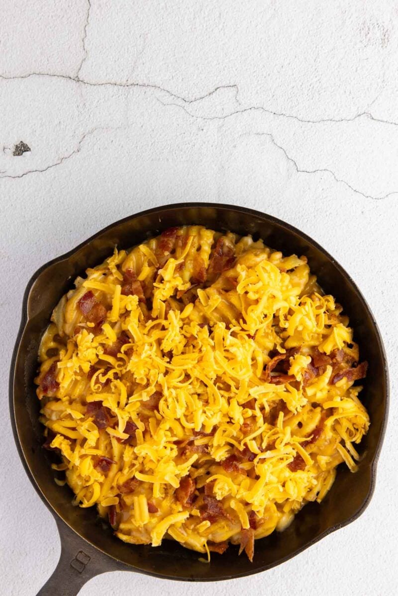 A cast iron skillet filled with macaroni and cheese topped with shredded cheddar cheese and bacon pieces, set on a light-colored surface.