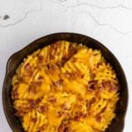 A cast iron skillet filled with rotini pasta baked in cheddar cheese sauce, topped with crispy bacon pieces, on a light textured surface.