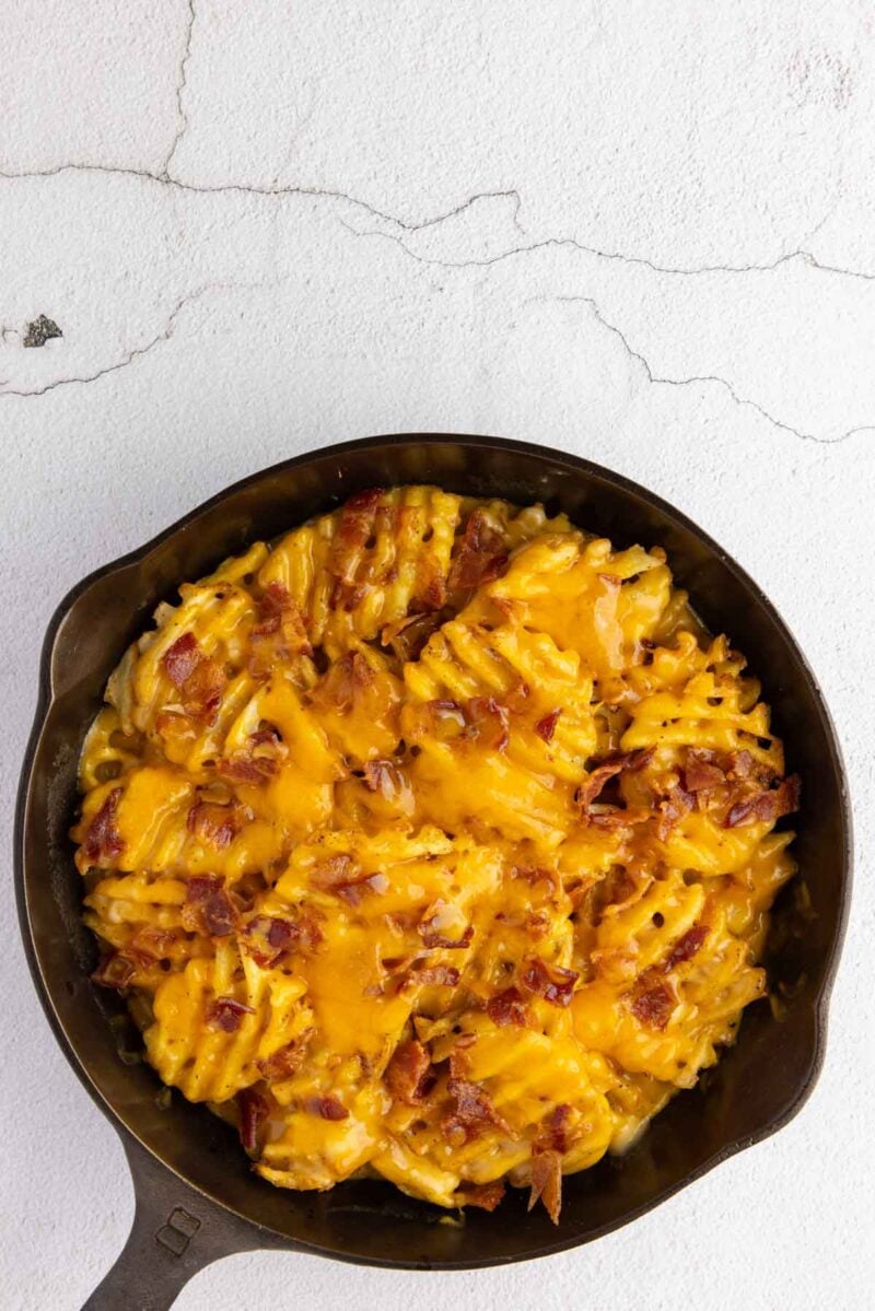 A cast iron skillet filled with rotini pasta baked in cheddar cheese sauce, topped with crispy bacon pieces, on a light textured surface.