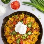 A skillet filled with cheesy potato nachos topped with sour cream, sliced jalapeños, chopped tomatoes, bacon, and green onions, surrounded by small bowls of toppings.