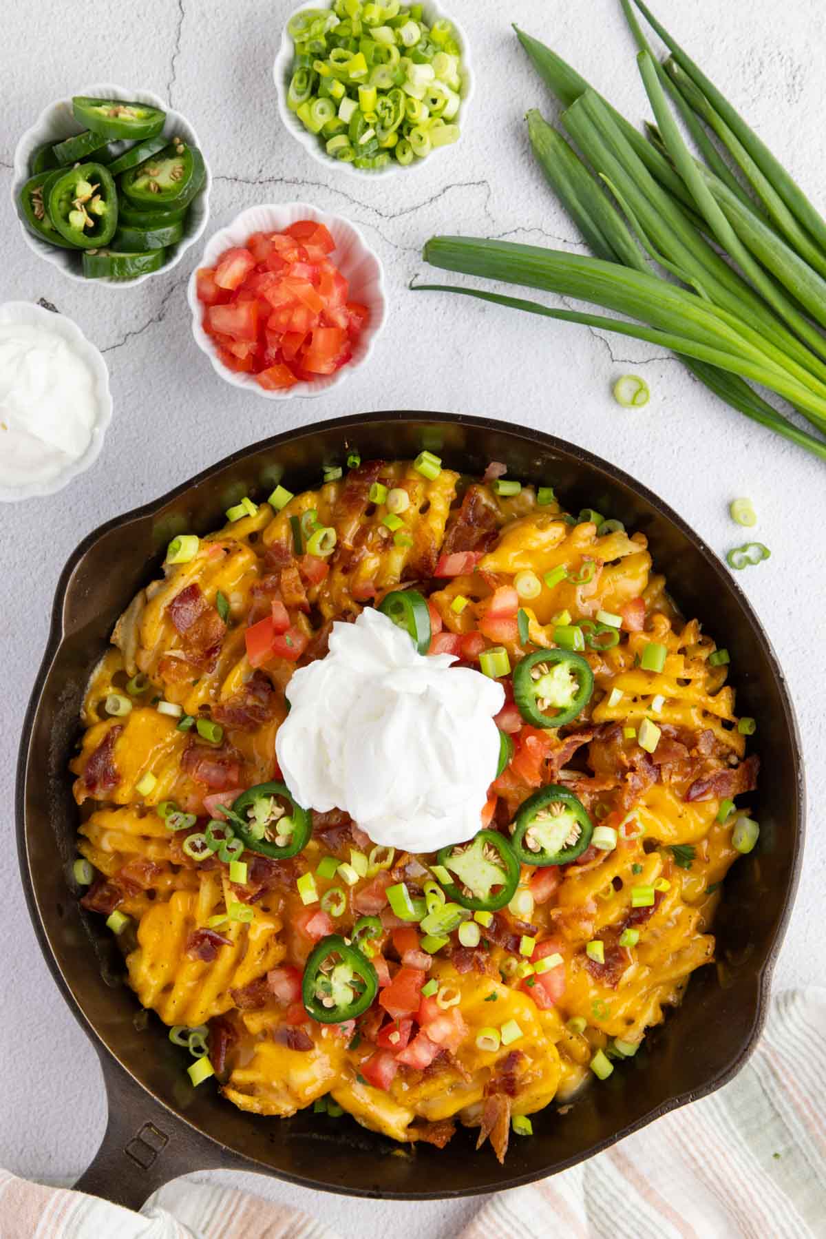 A skillet filled with cheesy potato nachos topped with sour cream, sliced jalapeños, chopped tomatoes, bacon, and green onions, surrounded by small bowls of toppings.