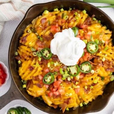 A skillet filled with waffle fries topped with melted cheese, bacon, jalapeños, diced tomatoes, green onions, and a dollop of sour cream.