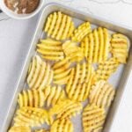 A baking tray with uncooked waffle-cut potato fries sprinkled with seasoning, next to a bowl containing more seasoning on a light textured surface.