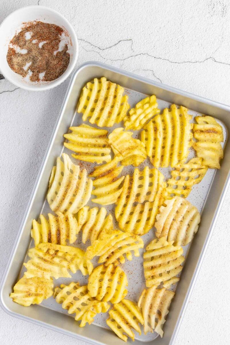 A baking tray with uncooked waffle-cut potato fries sprinkled with seasoning, next to a bowl containing more seasoning on a light textured surface.