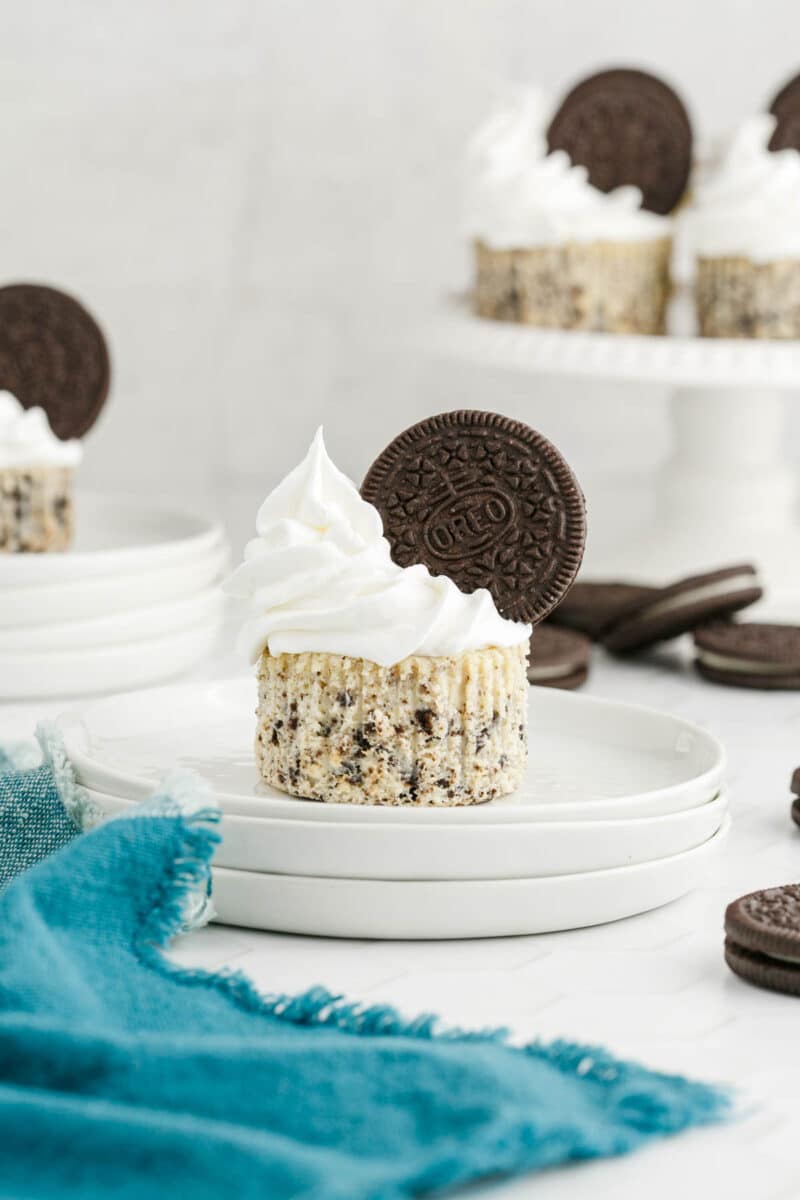 A cookies and cream cheesecake cupcake topped with whipped cream and an Oreo cookie sits on a white plate, with more cupcakes and Oreos in the background.