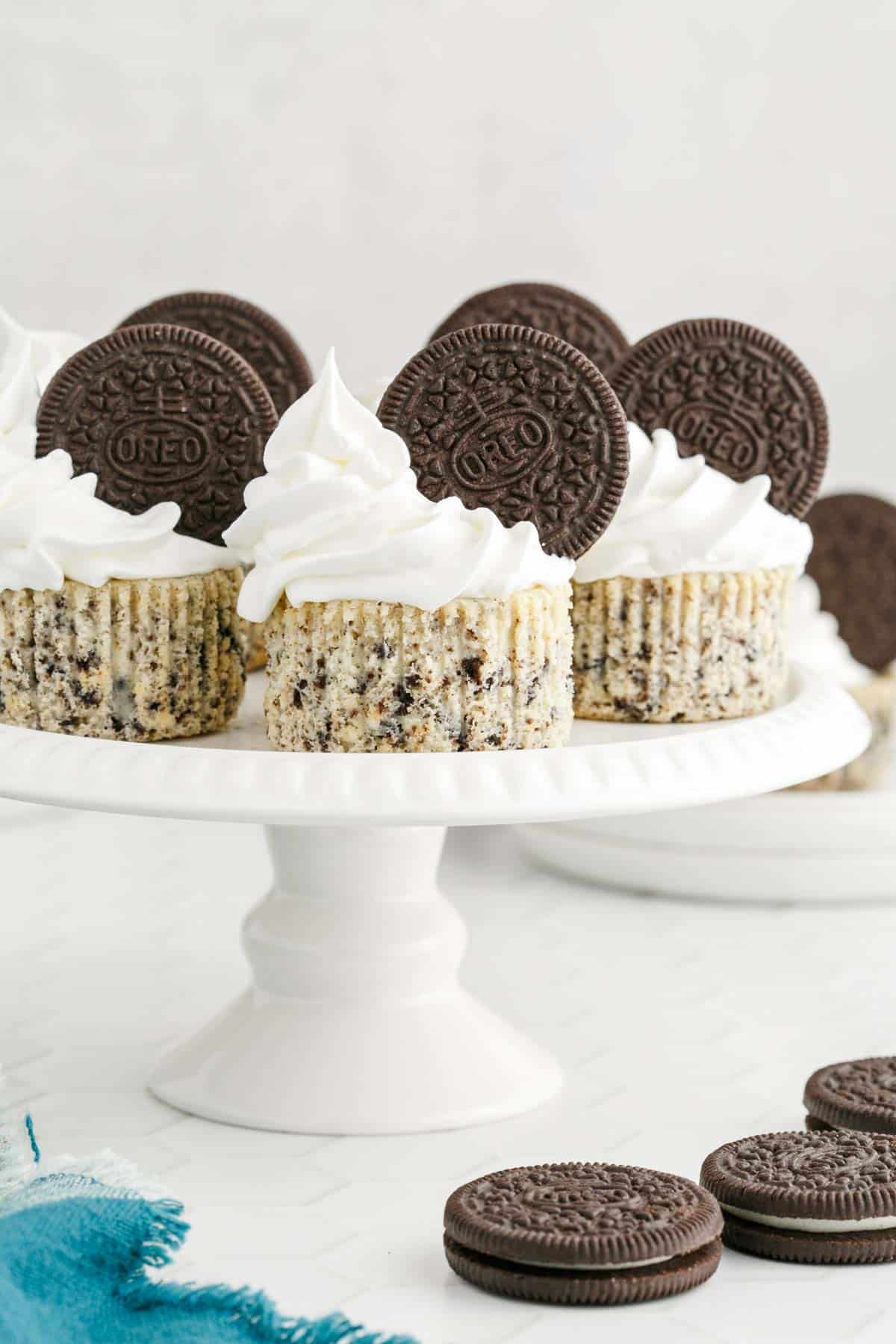 Mini Oreo cheesecakes with whipped cream and whole Oreo cookies on top are displayed on a white cake stand, with extra cookies and cheesecakes in the background.
