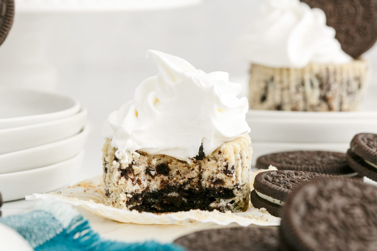 A cookies and cream cheesecake cupcake with whipped cream topping sits partially eaten among sandwich cookies on a white surface.