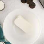 A block of cream cheese in a white mixing bowl, surrounded by Oreo cookies, sugar, a muffin tin, and a blue cloth on a white hexagon-tiled surface.