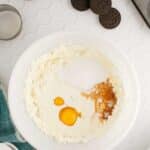 A mixing bowl with cream cheese, eggs, sugar, and vanilla, surrounded by Oreo cookies, a muffin pan with liners, a cup of milk, and a bowl of sugar on a white tiled surface.