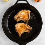Two seared pork chops in a black cast iron skillet, with bowls of diced potatoes and mixed vegetables on a white marble surface.