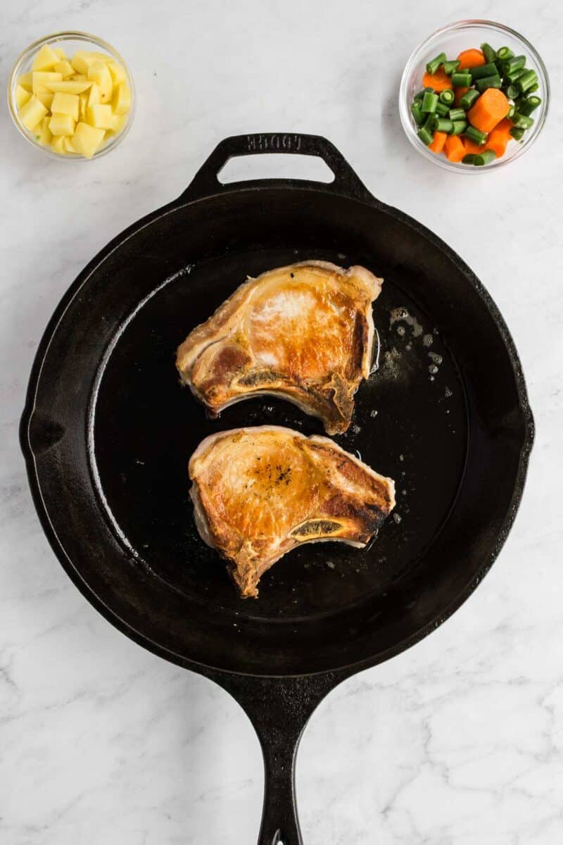 Two seared pork chops in a black cast iron skillet, with bowls of diced potatoes and mixed vegetables on a white marble surface.