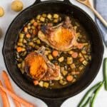 A cast iron skillet with two cooked pork chops on top of diced potatoes and carrots, surrounded by fresh carrots, green beans, and potatoes on a marble surface.