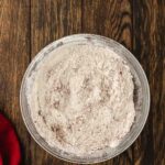 A glass bowl filled with a light brown dry flour mixture sits on a wooden surface next to a red cloth.